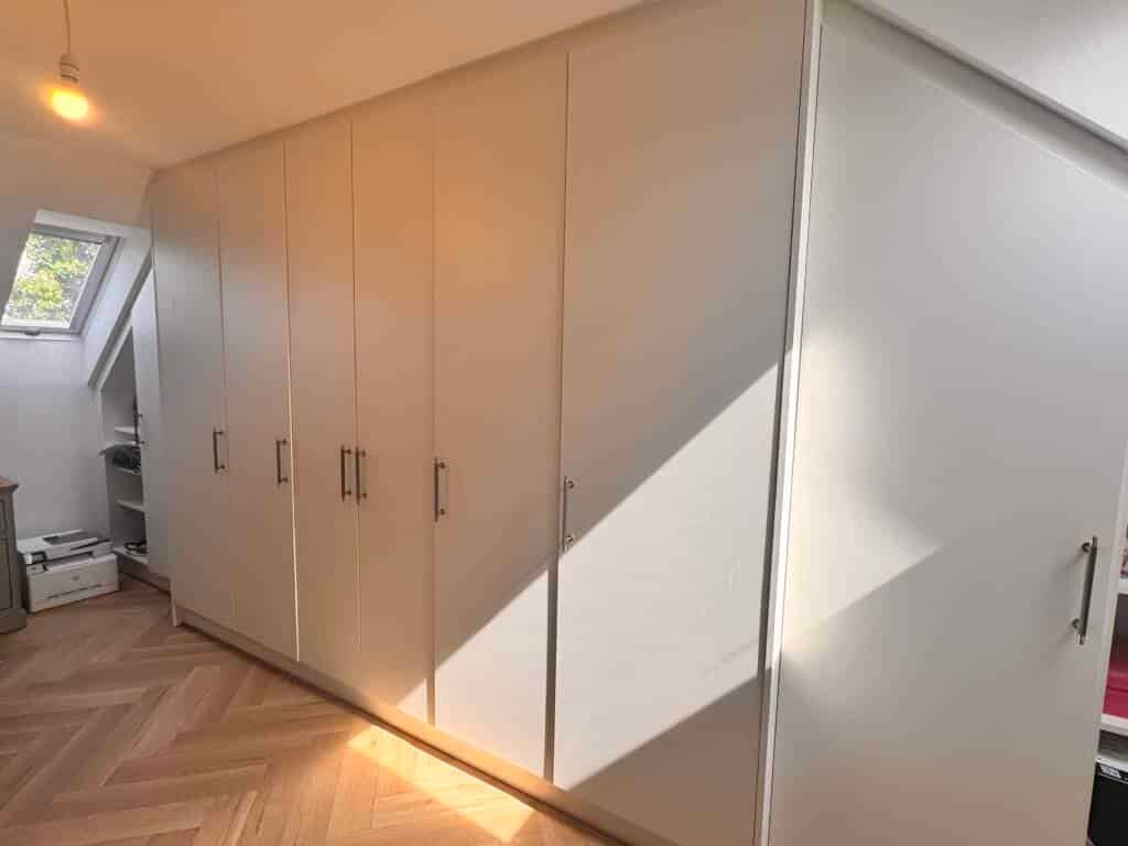 Expansive white built-in wardrobe design spanning a full wall in an attic room with light wood herringbone floors.