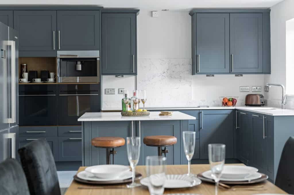 Sophisticated slate blue bespoke kitchen furniture featuring white marble countertops, a central kitchen island with leather stools, and integrated high-end appliances.
