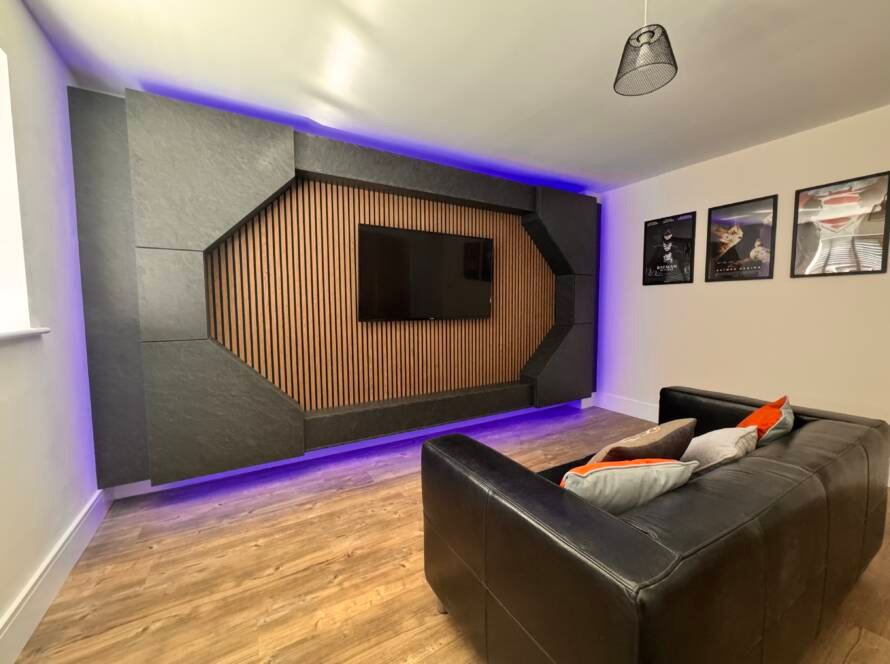A modern floating Media Wall with vertical oak wood slats and a dark charcoal geometric frame, illuminated by vibrant purple LED mood lighting in a home cinema setting.