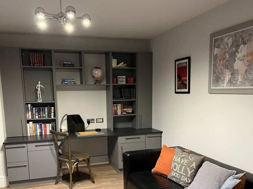 A cozy home office featuring dark grey fitted study furniture with a built-in desk, shelving unit, and a black leather sofa.